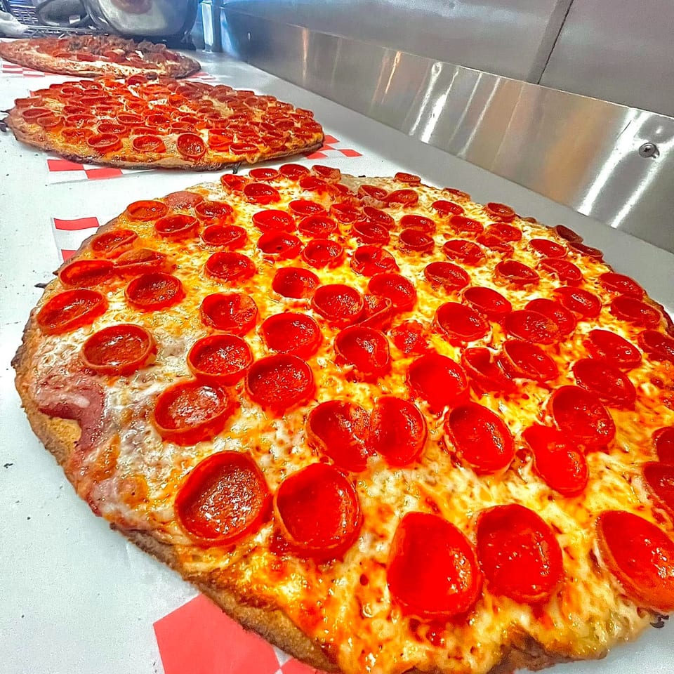 Several rectangular pepperoni pizzas with melted cheese sit on red-and-white checkered paper beside a commercial pizza oven.