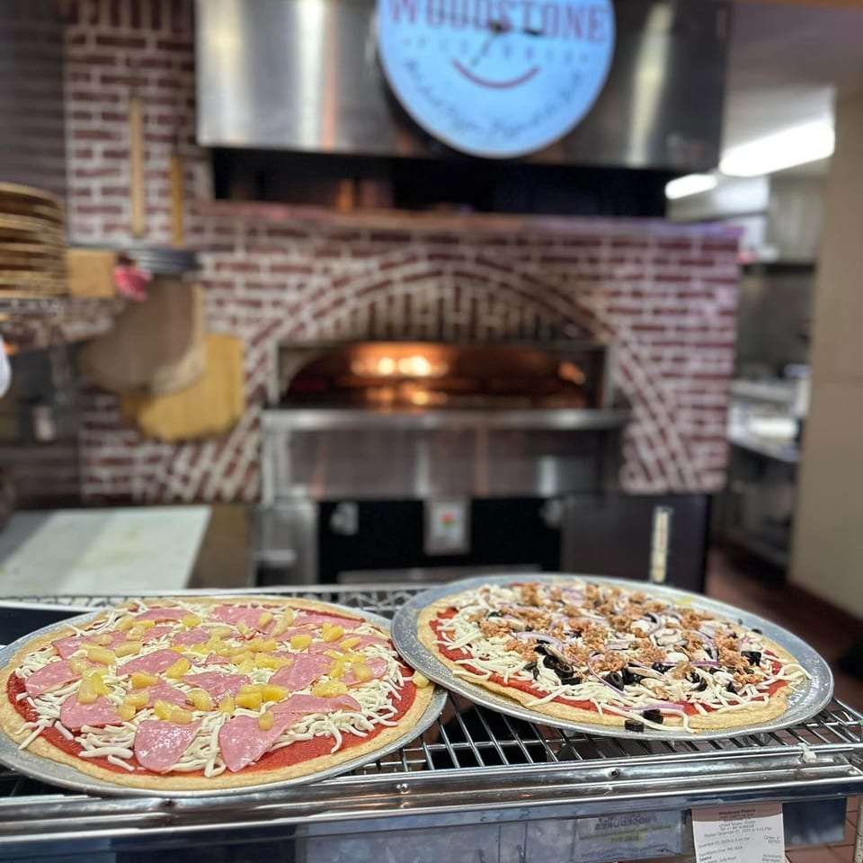 Two uncooked pizzas topped with salami, pineapple, vegetables, mushrooms, olives, and cheese sit before a brick pizza oven.