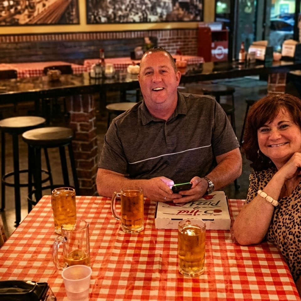 Three smiling people sit at a red and white checkered table with drinks and a pizza box.