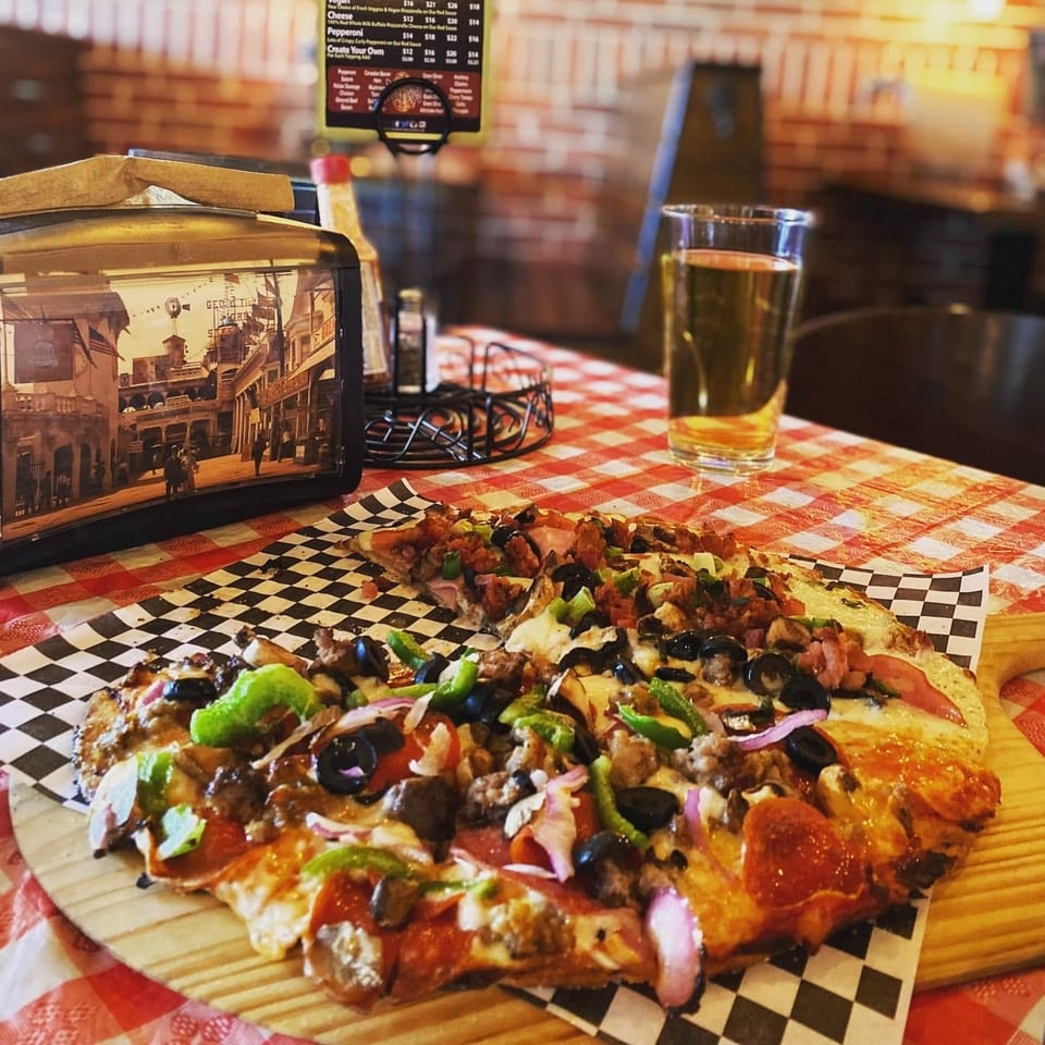 Loaded pizzas topped with pepperoni, olives, peppers, onions, meat, and cheese beside a pint of beer in a cozy restaurant.