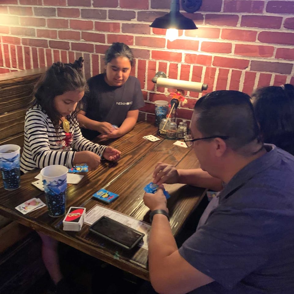 Group of diners gathered around a wooden restaurant table with drinks, playing cards, and a brick wall illuminated by sconces.