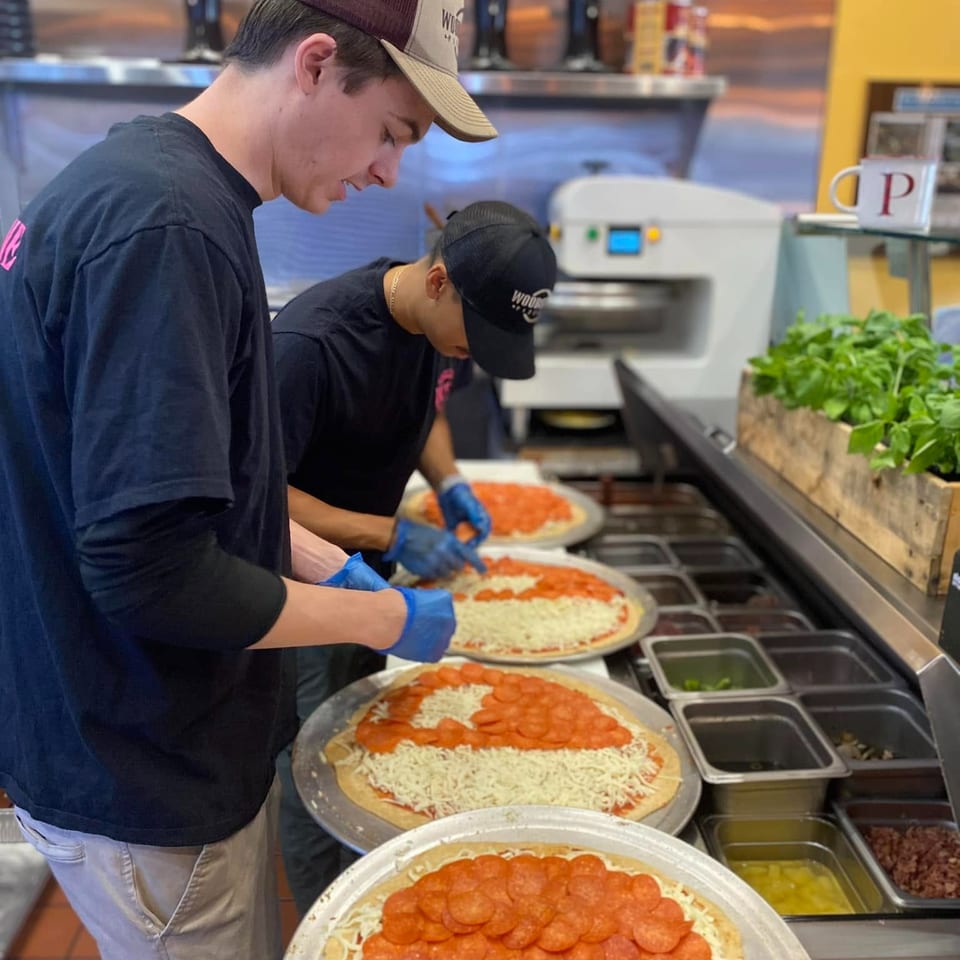 Restaurant staff in gloves prepare several pepperoni pizzas at a topping station.