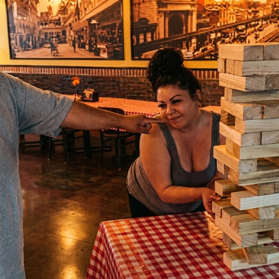 Two people playing a giant Jenga game on a red and white checkered table inside a restaurant.
